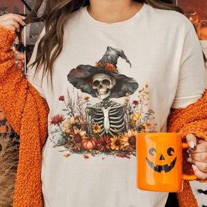May include: A white t-shirt with a skeleton wearing a witch hat and surrounded by fall foliage. The text "NATURAL" is on a black background in the lower left corner of the image.