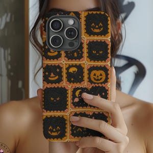 May include: Black and orange Halloween-themed phone case with a repeating pattern of jack-o'-lanterns, cats, and moons. The case has a textured appearance and a square design. The phone case is held in front of a person.