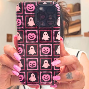 May include: Black phone case with a repeating pattern of pink jack-o'-lanterns and ghosts. The case has a square grid design with pink beaded borders. The phone case is held up in front of a mirror.