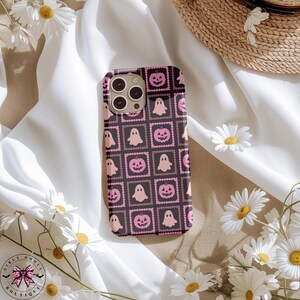 May include: Black phone case with a repeating pattern of pink ghosts and jack-o'-lanterns. The design is set within a square frame with a dotted border. The case is surrounded by white daisies and a straw hat.