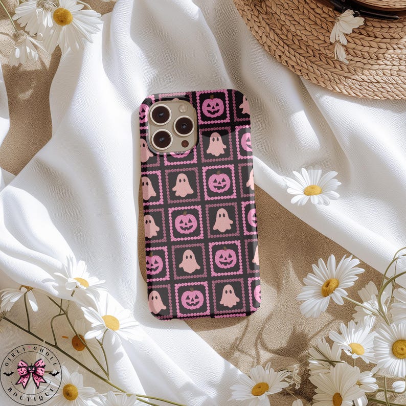 May include: Black phone case with a repeating pattern of pink ghosts and jack-o'-lanterns. The design is set within a square frame with a dotted border. The case is surrounded by white daisies and a straw hat.
