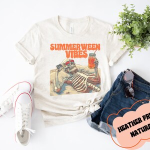 May include: A beige t-shirt with the text "Summerween Vibes" in orange and red. A skeleton wearing sunglasses is lounging on a beach with a glass of orange juice. The text "Heather Prism Natural" is written in a cloud shape.