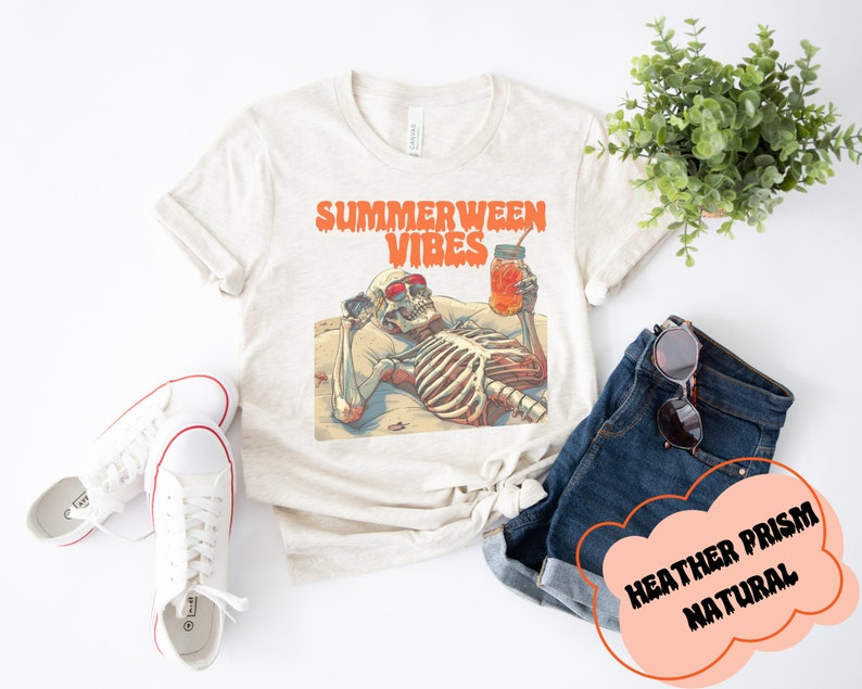 May include: A beige t-shirt with the text "Summerween Vibes" in orange and red. A skeleton wearing sunglasses is lounging on a beach with a glass of orange juice. The text "Heather Prism Natural" is written in a cloud shape.