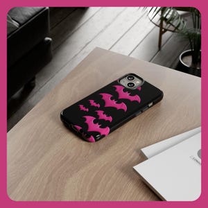May include: A black phone case featuring a pink bat design. The case is made from hard plastic and has a raised edge to protect the screen.