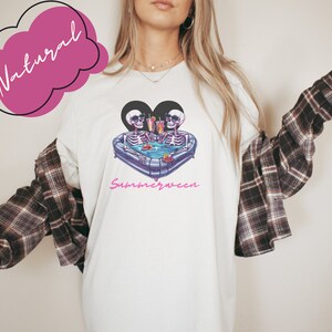 May include: White t-shirt with a graphic of two skeletons in a heart-shaped inflatable pool with drinks. The text "Summerween" is printed below the graphic.