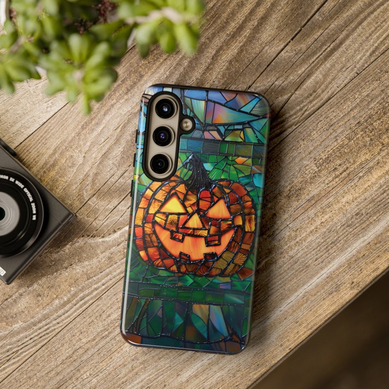 May include: A stained glass style phone case with a jack-o-lantern design. The case is made of a clear material with a colorful stained glass pattern. The jack-o-lantern is orange and has a black outline. The case has a camera cutout on the back.