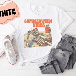 May include: White t-shirt with a graphic of a skeleton wearing sunglasses and holding a glass of orange juice. The text "Summerween Vibes" is printed on the shirt.