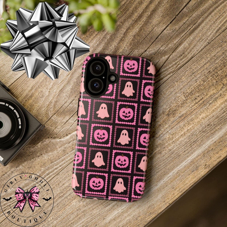 May include: A black and pink Halloween-themed phone case featuring a repeating pattern of pink jack-o'-lanterns and ghosts within pink square frames. The case has a camera cutout and is on a wooden surface. The Girly Ghoul Boutique logo is visible.