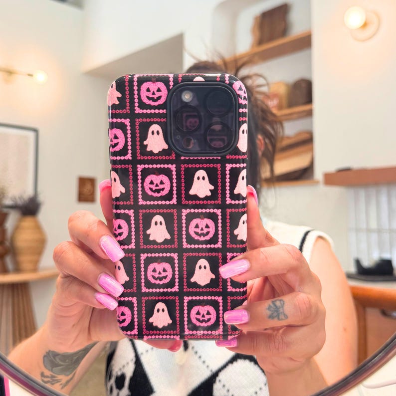 May include: Black phone case with a repeating pattern of pink jack-o'-lanterns and ghosts. The case has a square grid design with pink beaded borders. The phone case is held up in front of a mirror.