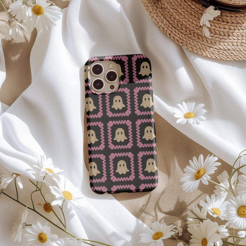 K&ouml;nnte beinhalten: Schwarze Smartphone-H&uuml;lle mit einem sich wiederholenden Muster aus beigen Geistern in rosa Zickzackrahmen. Die H&uuml;lle hat eine matte Oberfl&auml;che und einen Kameraausschnitt. Das Design befindet sich auf einem wei&szlig;en Stoffhintergrund mit G&auml;nsebl&uuml;mchen und einem Strohhut.