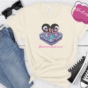 May include: A white t-shirt with a graphic of two skeletons in a heart-shaped inflatable pool with drinks. The text "Summerween" is printed below the graphic.