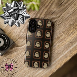Może przedstawiać: Czarne etui na telefon z powtarzającym się wzorem beżowych postaci duch&oacute;w w brązowych kwadratowych ramkach. Etui leży na drewnianej powierzchni, obok srebrnej kokardy i aparatu. Widoczne jest logo "Girly Ghost Boutique".