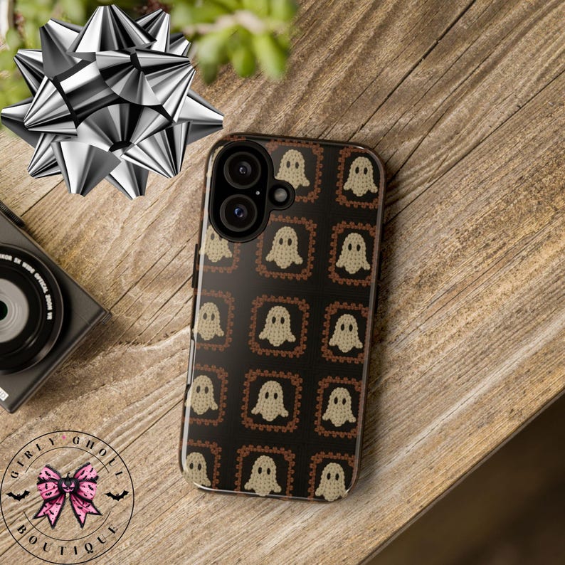 Może przedstawiać: Czarne etui na telefon z powtarzającym się wzorem beżowych postaci duch&oacute;w w brązowych kwadratowych ramkach. Etui leży na drewnianej powierzchni, obok srebrnej kokardy i aparatu. Widoczne jest logo "Girly Ghost Boutique".