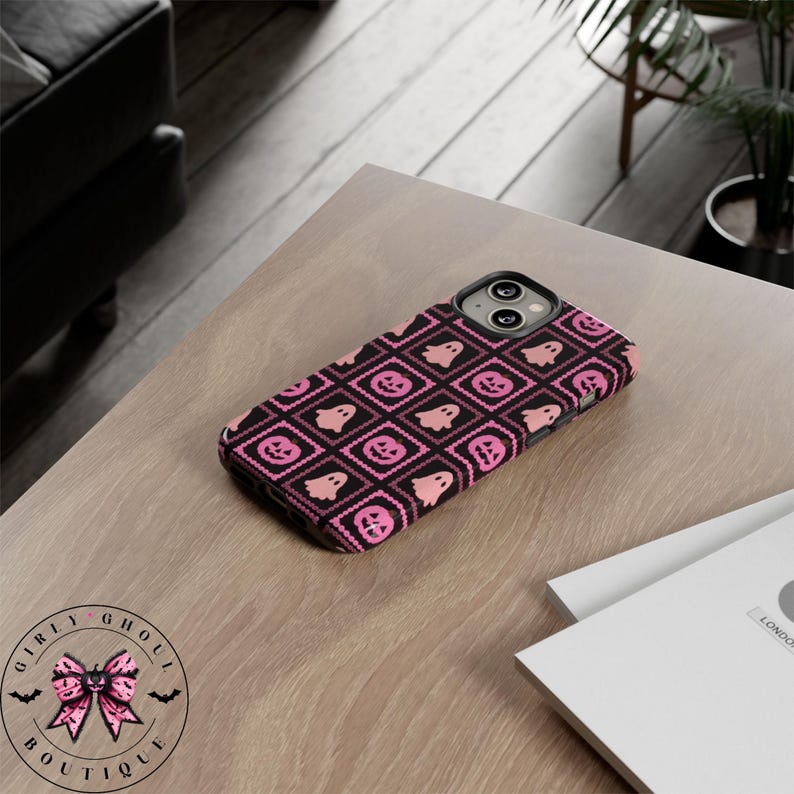 May include: Black phone case with a pink Halloween pattern featuring ghosts and jack-o'-lanterns. The case is on a light wood surface. The case has a black camera lens area. The case is from Girly Ghoul Boutique.
