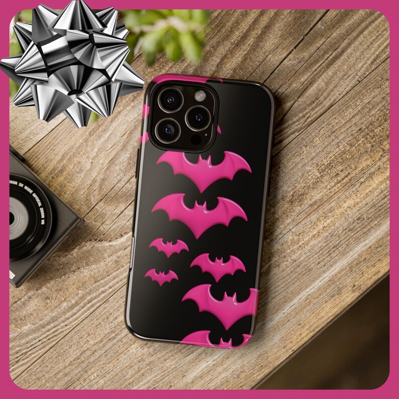 May include: Black phone case with a pink bat pattern. The case has a camera lens cutout and a black background.