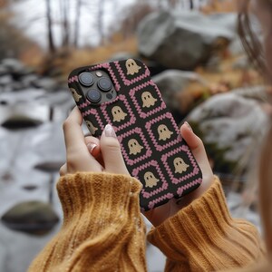 K&ouml;nnte beinhalten: Schwarze Smartphone-H&uuml;lle mit einem sich wiederholenden Muster aus beigen Geisterfiguren in rosa und schwarzen Quadraten. Die H&uuml;lle wird in den H&auml;nden einer Person gehalten, mit einem verschwommenen Hintergrund aus Bach und Bl&auml;ttern.