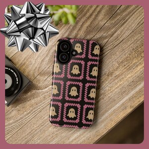 K&ouml;nnte beinhalten: Eine schwarze Smartphone-H&uuml;lle mit einem sich wiederholenden Muster aus beigen Geistern in rosa Zickzackrahmen. Die H&uuml;lle liegt auf einer Holzoberfl&auml;che, mit einer silbernen Schleife und einer Kamera im Hintergrund.