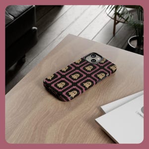 K&ouml;nnte beinhalten: Eine schwarze Smartphone-H&uuml;lle mit einem sich wiederholenden Muster aus beigefarbenen Geistern in rosa und schwarzen Quadraten. Die H&uuml;lle liegt auf einer hellen Holzoberfl&auml;che, mit einem wei&szlig;en Notizblock und einer Pflanze im Hintergrund.