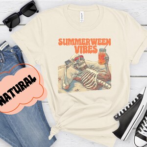 May include: A beige t-shirt with a graphic of a skeleton wearing sunglasses and holding a glass of iced tea. The text "Summerween Vibes" is printed in orange above the skeleton.