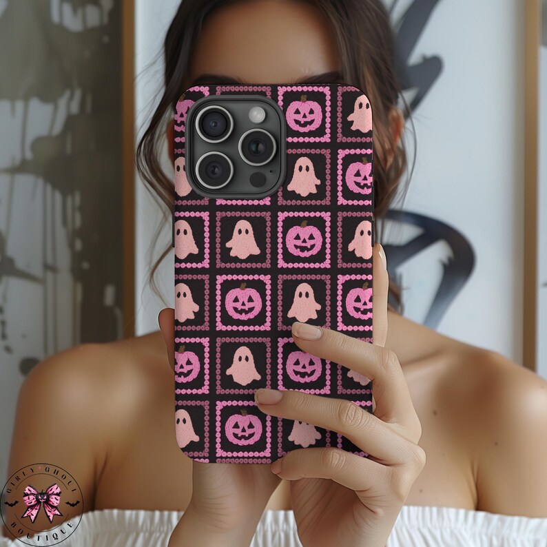 May include: Black phone case with a repeating pattern of pink ghosts and jack-o'-lanterns. The design is set within pink square frames on a black background. The phone case is held in front of a person.