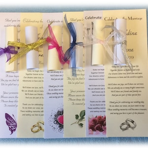 May include: Four wedding favor scrolls tied with ribbon and a card with the text "Celebrate the Marriage of [Name] and [Name]" and the date "2019". Each scroll has a different color ribbon and a different design on the card. The cards are all cream colored with a floral design.