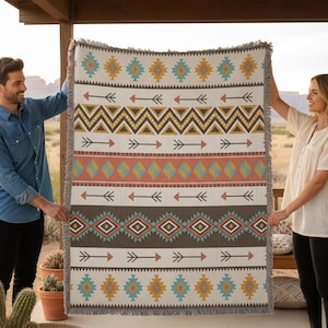 May include: A woven blanket with a Southwestern design. The blanket features geometric patterns in shades of brown, orange, teal, and yellow on a cream background. Arrows and diamond shapes are prominent. The blanket has a fringed edge.