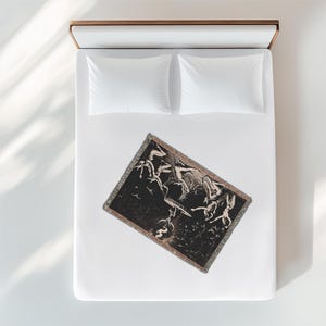 A black and white tapestry with a design of figures in mid-air, falling from the sky. The tapestry is on a white bed with white pillows.