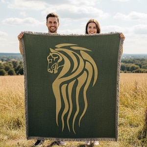 Op de afbeelding: Een donkergroene sprei met een gouden paardenkopontwerp. De paardenkop is gestileerd met vloeiende lijnen. De deken heeft een gefranjerde rand en wordt door twee mensen in een veld omhoog gehouden.