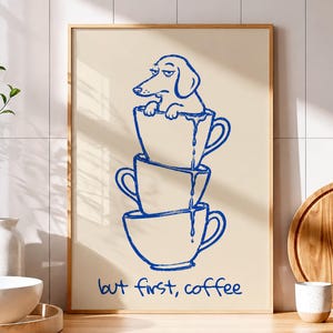 May include: A rectangular wall art print in a light wooden frame featuring a blue line drawing of a dog sitting inside a stack of three coffee cups, with the text "but first, coffee" written in blue script at the bottom of the beige background.