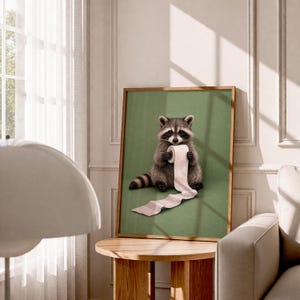 May include: A framed art print featuring a raccoon holding a roll of toilet paper against a green background. The raccoon is depicted in detail with gray and brown fur. The artwork is displayed in a room with a wooden table and a window.