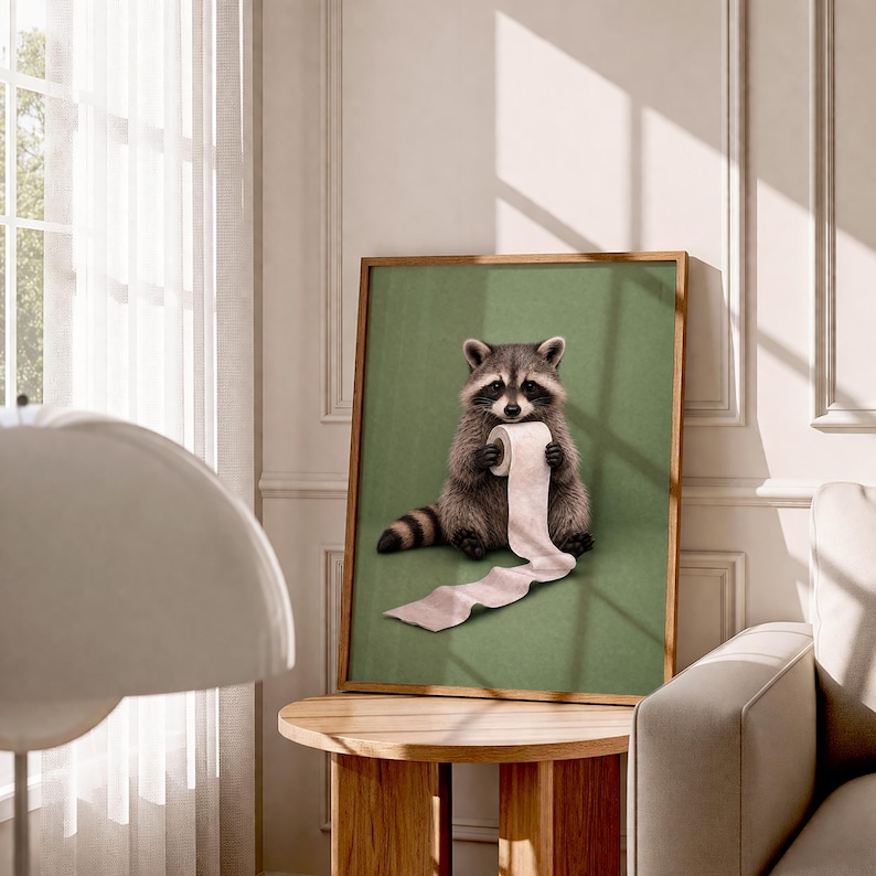 May include: A framed art print featuring a raccoon holding a roll of toilet paper against a green background. The raccoon is depicted in detail with gray and brown fur. The artwork is displayed in a room with a wooden table and a window.
