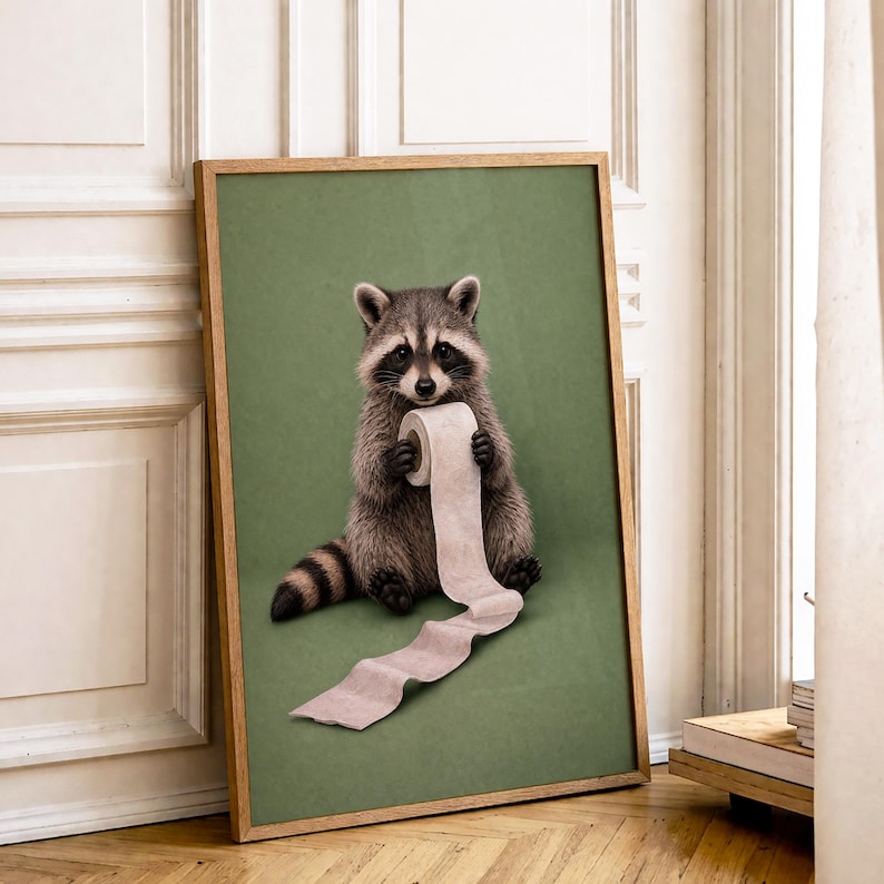 May include: A framed print featuring a raccoon holding a roll of toilet paper against a green background. The raccoon is depicted in realistic detail, with gray and brown fur and a black mask. The frame is a light brown color.