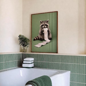 May include: A framed print featuring a raccoon holding a roll of toilet paper against a green background. The artwork has a brown frame and is displayed in a bathroom setting with green and white accents. The raccoon is depicted in a realistic style.