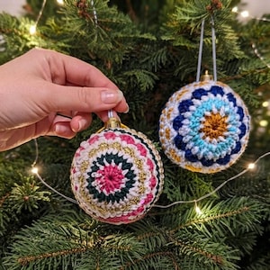 May include: Two spherical Christmas ornaments featuring intricate crochet patterns. One ornament displays concentric rings of pink, dark green, white, and gold yarn. The second ornament features a blue, light blue, white, and gold floral-like design.