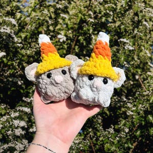 May include: Two small, plush, crocheted mouse figures, one light gray and one white, each wearing a pointed hat colored in horizontal stripes of white, orange, and yellow, resembling candy corn, with black bead eyes and small pink noses.