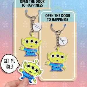 May include: Two keychains featuring a green alien character with three eyes and a blue jumpsuit. The keychains have silver rings and charms with the letters "L" and "A". The packaging has the text "Open the door to happiness" and "Get me free!"
