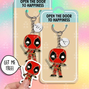 Op de afbeelding: Een rode en zwarte Deadpool-sleutelhanger met een zilveren sleutelring en een bedel met de tekst "Open de deur naar geluk". De sleutelhanger wordt getoond op een witte kaart met dezelfde tekst. De kaart heeft ook een sticker van Deadpool met de tekst "Haal me gratis!"