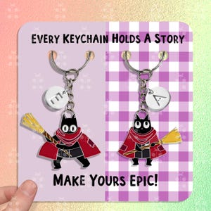 Può includere: Due ciondoli portachiavi a forma di gatto nero con mantelli rossi e scope. I ciondoli sono attaccati a portachiavi argentati con ciondoli a forma di lettera iniziale. Lo sfondo è un motivo a scacchi bianco e viola con il testo "EVERY KEYCHAIN HOLDS A STORY" e "MAKE YOURS EPIC!"