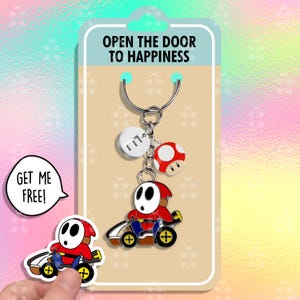 Peut inclure: Un porte-clés avec un personnage Shy Guy rouge dans un kart, un champignon rouge et blanc, et un disque blanc avec la lettre "E". Le porte-clés est attaché à une carte qui dit "OUVREZ LA PORTE AU BONHEUR". Un autocollant dit "OBTENEZ-MOI GRATUITEMENT!"