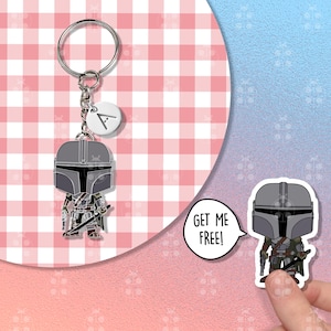 Puede incluir: Un llavero de Mandaloriano con un dise&ntilde;o de personaje estilizado. El llavero tiene un anillo plateado y un peque&ntilde;o dije blanco con la letra "A". La figura del Mandaloriano es en tonos grises, verdes y marrones. El fondo es un patr&oacute;n de cuadros rosas y blancos.