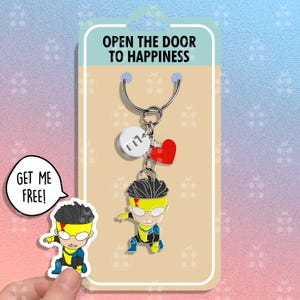 May include: A yellow and blue superhero keychain with a red heart charm and a silver charm with the letter "M" on it. The keychain is attached to a silver key ring. The keychain is on a white card with the text "Open the door to happiness" and a cartoon image of the superhero. The card also has a speech bubble that says "Get me free!"