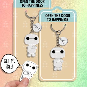 May include: Two silver Baymax keychain charms with a heart on the front. The charms are attached to silver key rings with a small silver charm with a letter on it. The keychains are displayed on a beige card with the text "Open the door to happiness".