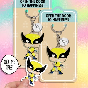 Op de afbeelding: Twee sleutelhangers met de tekst "Open the door to happiness" en een geel en zwart cartoonfiguur. De sleutelhangers hebben een zilveren hanger met de letters "L" en "A" erop. De afbeelding bevat ook een sticker van hetzelfde cartoonfiguur met de tekst "Get me free!"