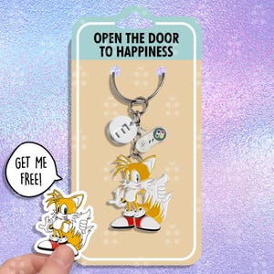 May include: A keychain featuring a cartoon character with a yellow and orange tail, a red and white shirt, and blue shoes. The keychain also includes a silver charm with the letter "M" and a silver charm shaped like a video game controller. The text "Open the door to happiness" is printed on the card behind the keychain. The text "Get me free!" is printed on a separate card with a cartoon character with a yellow and orange tail.
