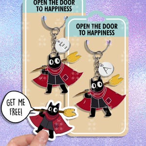 Puede incluir: Dos llaveros de gato negro con capas rojas y escobas. Los llaveros tienen un dije plateado con las letras "E" y "A" en ellos. El texto "Abre la puerta a la felicidad" está impreso en el empaque. Una calcomanía con un gato negro sosteniendo una escoba y el texto "¡Libérame!" se muestra en primer plano.