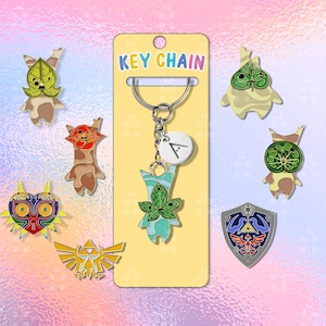 Puede incluir: Un llavero con un dije de personaje verde y azul, un dije blanco con la letra "A" y varios pines de personajes. El llavero está en una tarjeta amarilla que dice "KEY CHAIN" en colores del arcoíris. Otros pines incluyen un escudo y un emblema dorado.