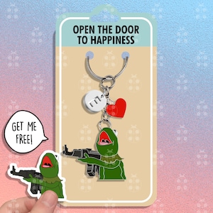 May include: A green frog holding a gun with the text "Get me free!" in a speech bubble. The frog is on a keychain with a silver ring, a red heart charm, and a silver charm with the letter "M". The keychain is on a card with the text "Open the door to happiness".