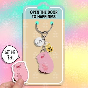 Könnte beinhalten: Ein rosa Schweine-Schlüsselanhänger mit einem silbernen Schlüsselring und Anhängern. Die Anhänger umfassen einen silbernen Kreis mit dem Buchstaben "M" und eine gelbe Hand mit der Nummer "3". Der Schlüsselanhänger ist auf einer Karte verpackt, auf der "Öffne die Tür zum Glück" steht.