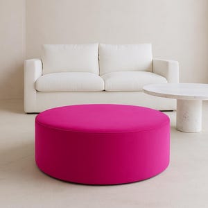 May include: A large, round, vibrant fuchsia ottoman with a smooth, solid-colored fabric surface. The piece features a low-profile, cylindrical shape that sits directly on the floor, providing a bold pop of color in a neutral-toned room.