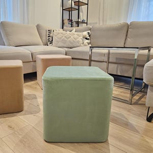 Puede incluir: Escena de salón con un sofá seccional beige, una mesa auxiliar de metal con tapa de cristal y tres otomanas cuadradas en tonos beige, melocotón y verde. Las otomanas miden aproximadamente 45 cm de ancho.